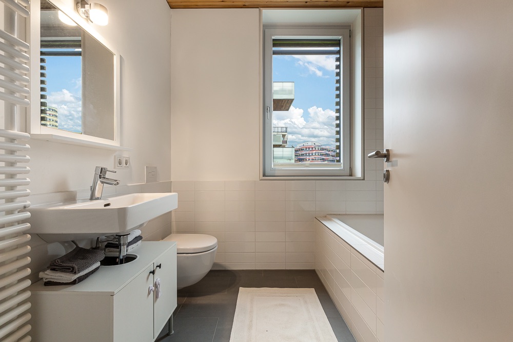 Helles Badezimmer mit Waschbecken, Toilette, Badewanne und Fenster mit Blick nach draußen.
