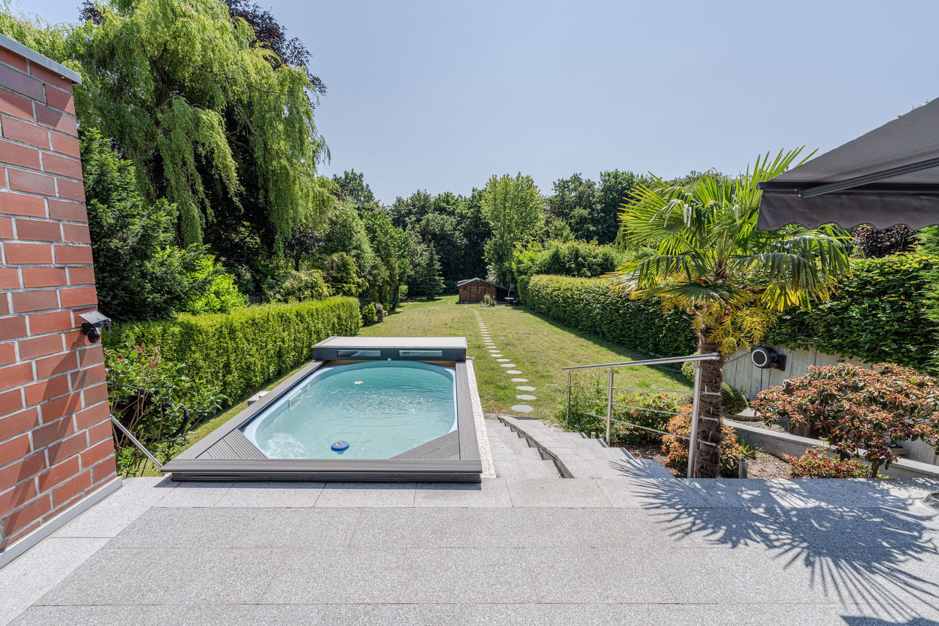 Terrasse mit Pool, Treppe zum Garten, Rasenfläche und Gartenhaus im Hintergrund.