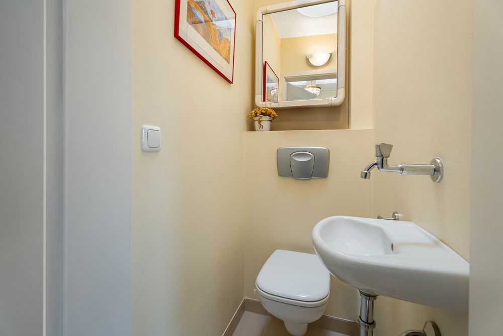 Kleines Badezimmer mit Wandtoilette, kleinem Waschbecken, Spiegel und Bild an der Wand.