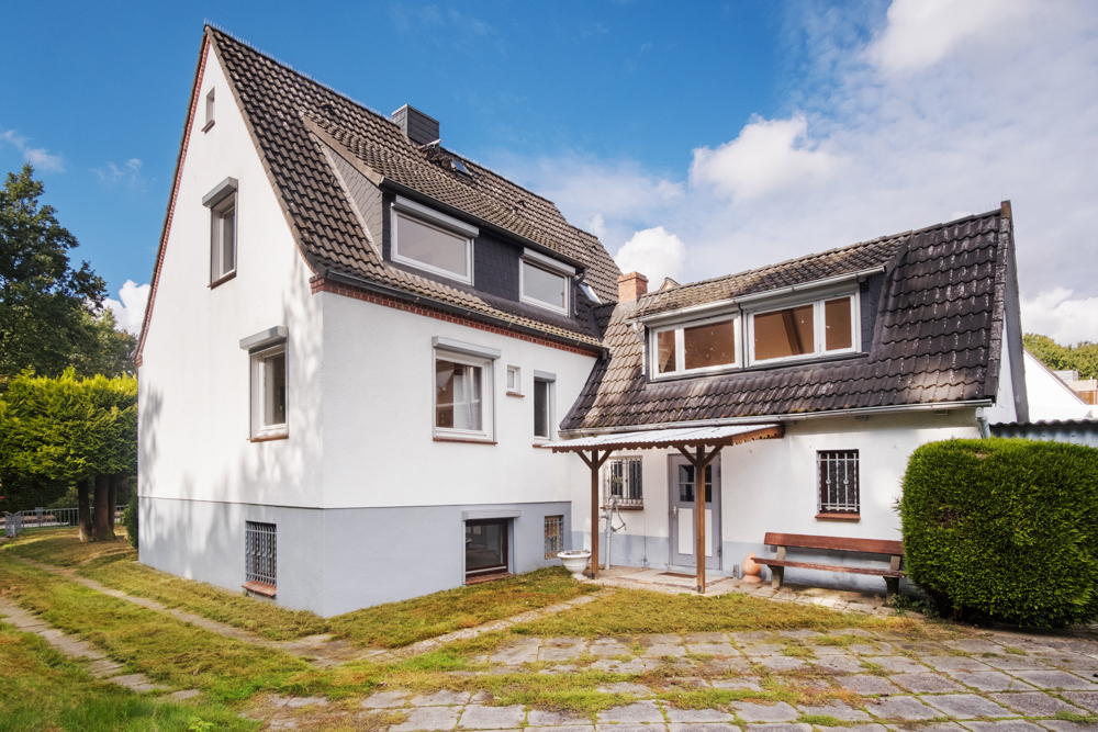 Weißes Wohnhaus mit dunklem Satteldach, mehreren Fenstern und kleinem Vordach im Garten.