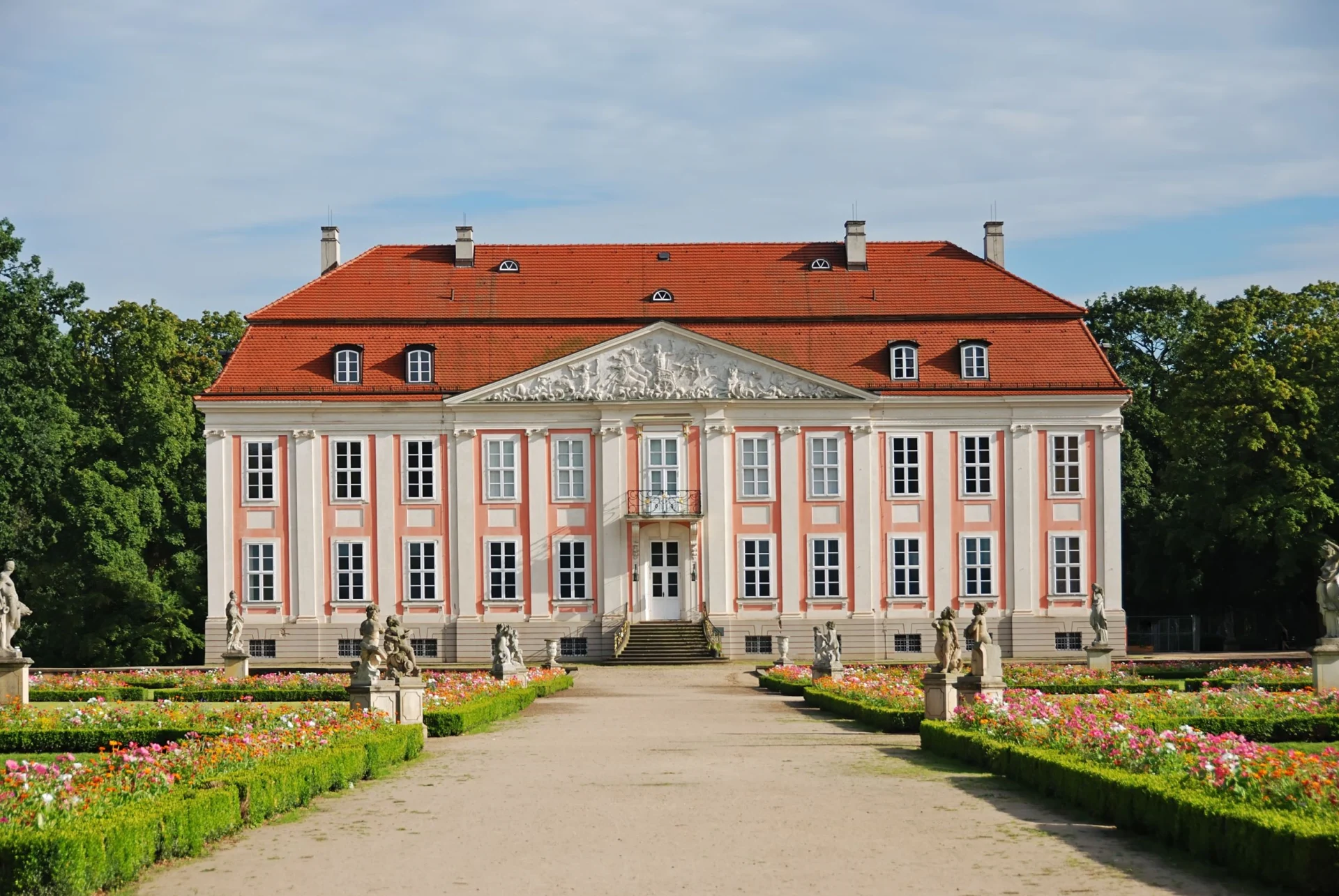 Großes Herrenhaus mit rotem Dach und gepflegtem Garten mit Blumen und Statuen davor.