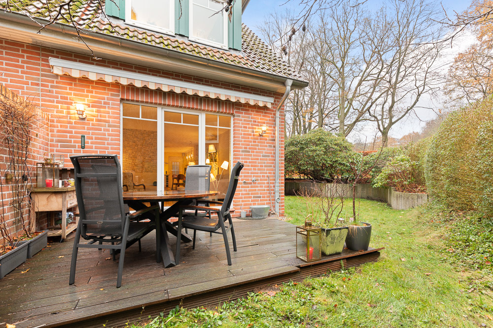 Holzterrasse mit Tisch und Stühlen an einem Backsteinhaus, daneben Garten mit Rasen und Sträuchern.