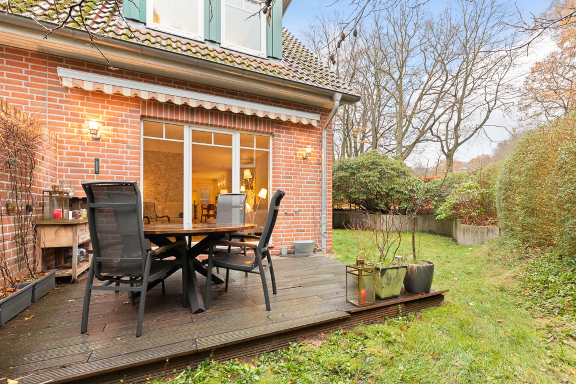 Holzterrasse mit Tisch und Stühlen an einem Backsteinhaus, daneben Garten mit Rasen und Sträuchern.