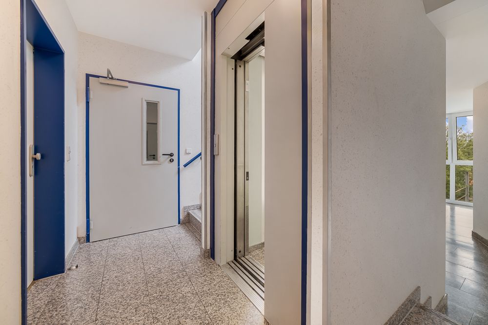 Flur mit geöffnetem Aufzug, Tür und Treppenaufgang, Tageslicht durch Fenster.