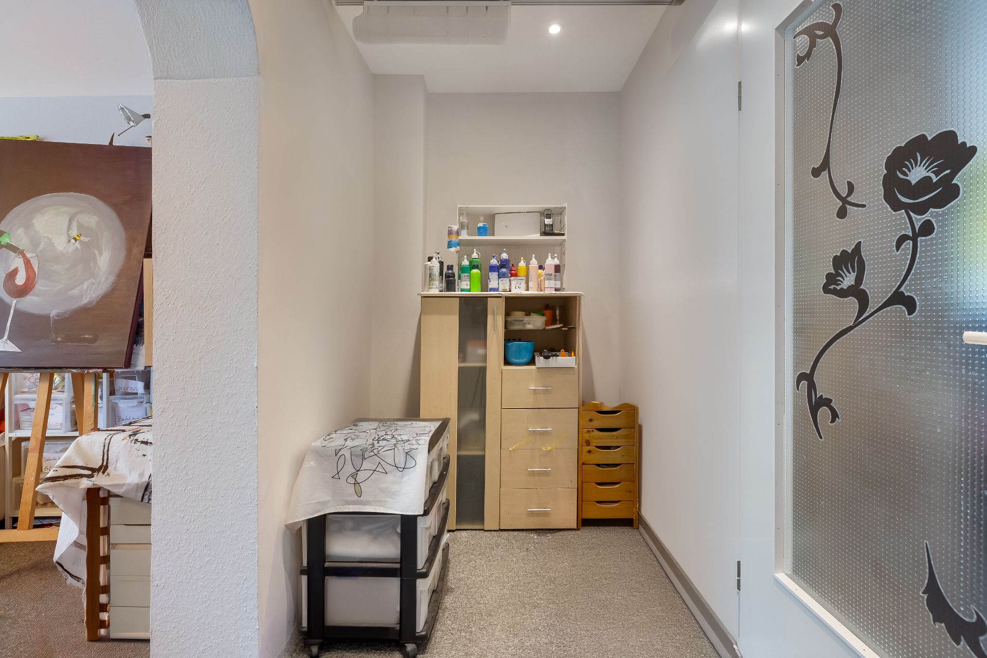 Kleiner Raum mit Schrank, Regalen und Malutensilien, Tür mit Blumenmuster rechts.