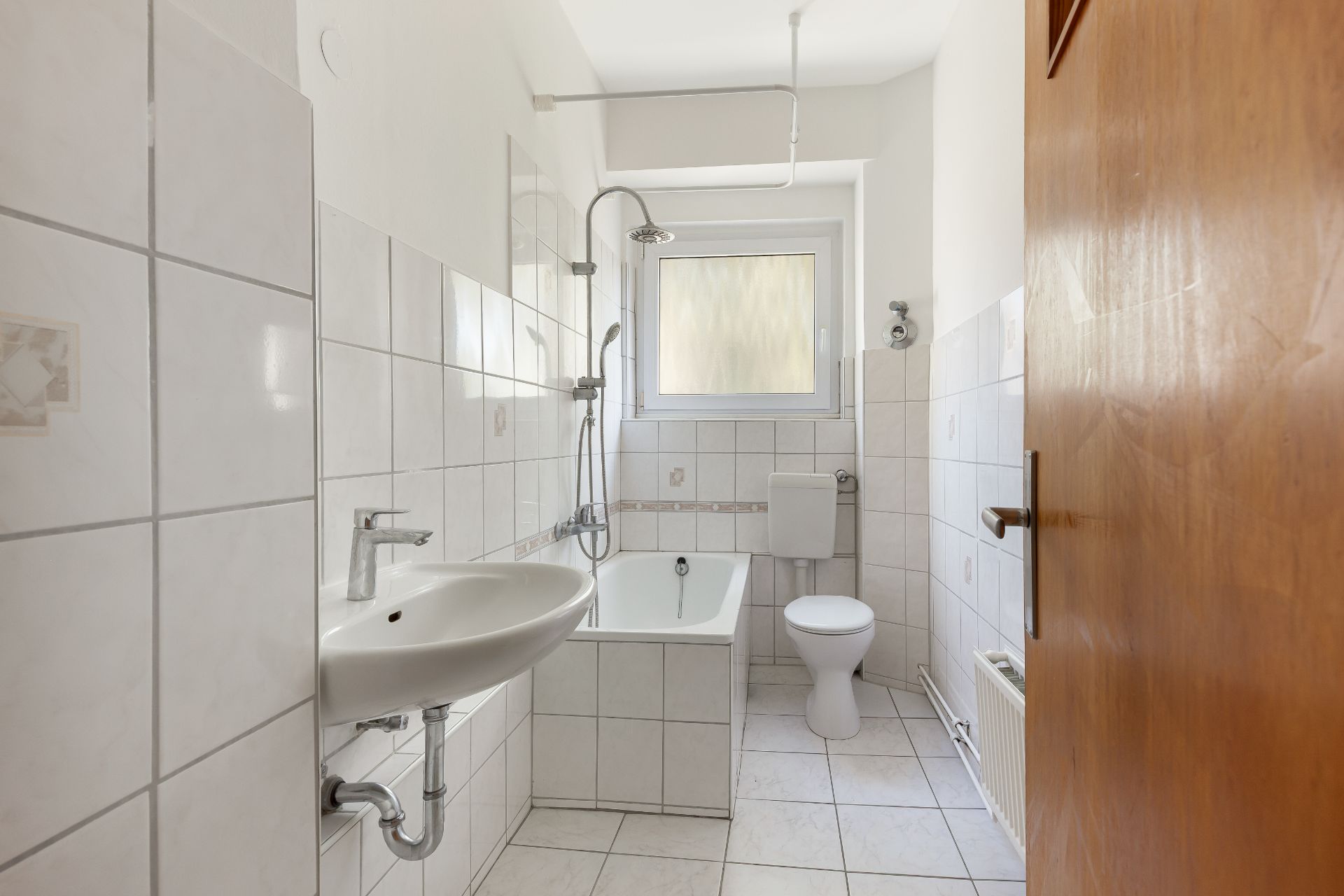 Helles Badezimmer mit Badewanne, Waschbecken, WC und Fenster, weiß gefliest.