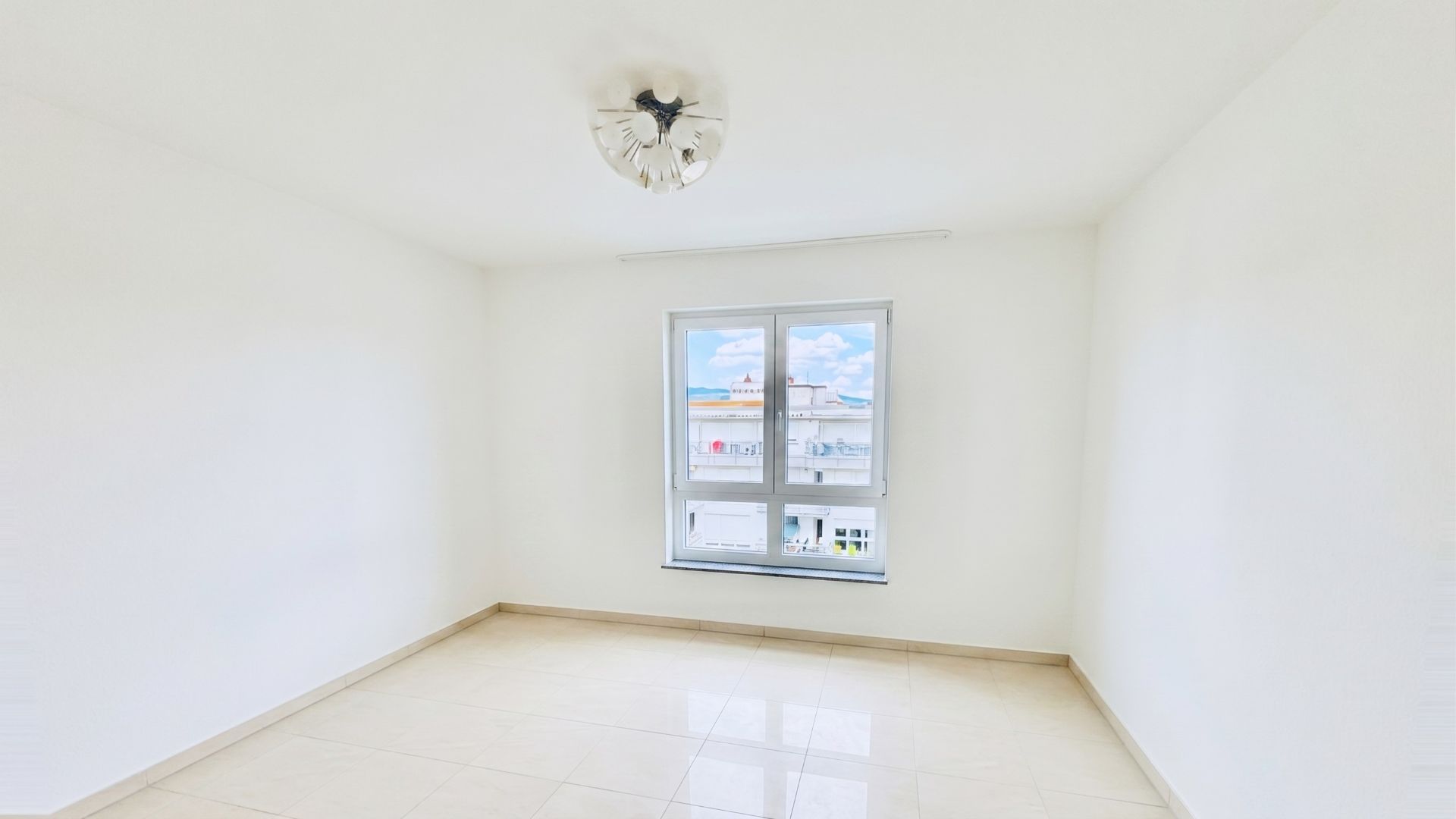 Helles, leeres Zimmer mit weißem Boden, weißer Decke und einem Fenster mit Blick nach draußen.