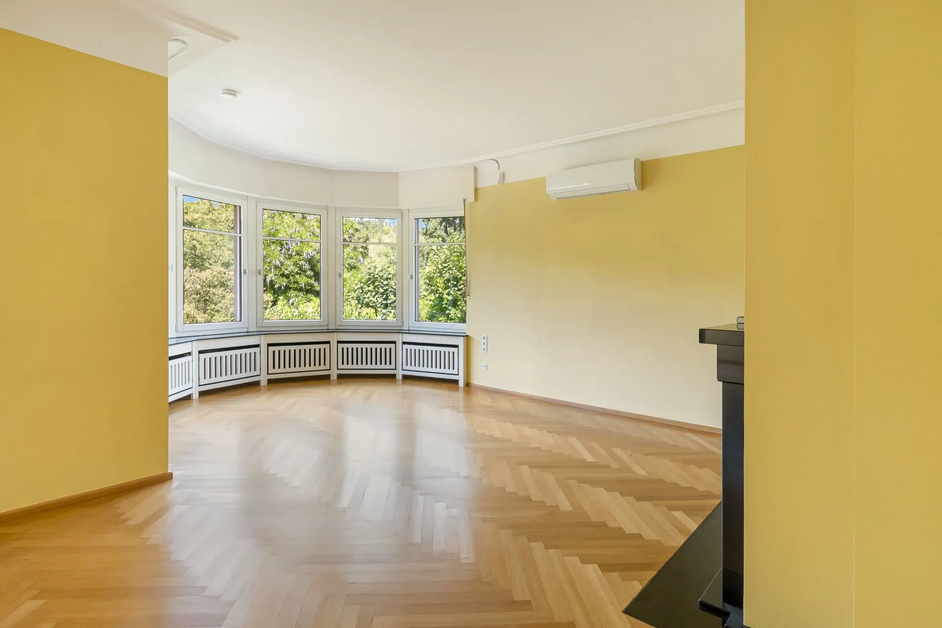 Helles Wohnzimmer mit großen Fenstern, Parkettboden und Klimaanlage an der Wand.