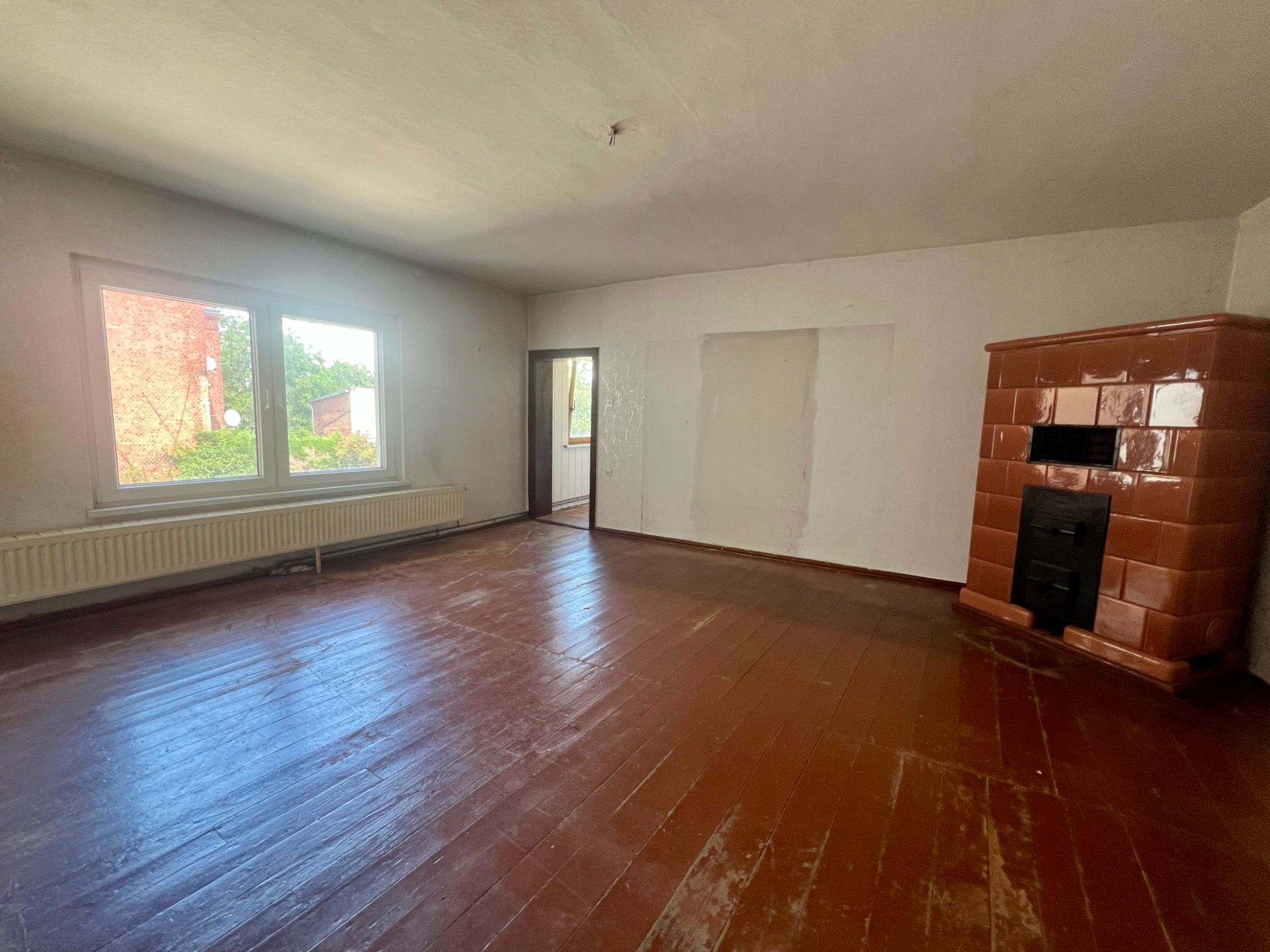 Leeres Zimmer mit braunem Holzboden, großem Fenster und braunem Kachelofen.