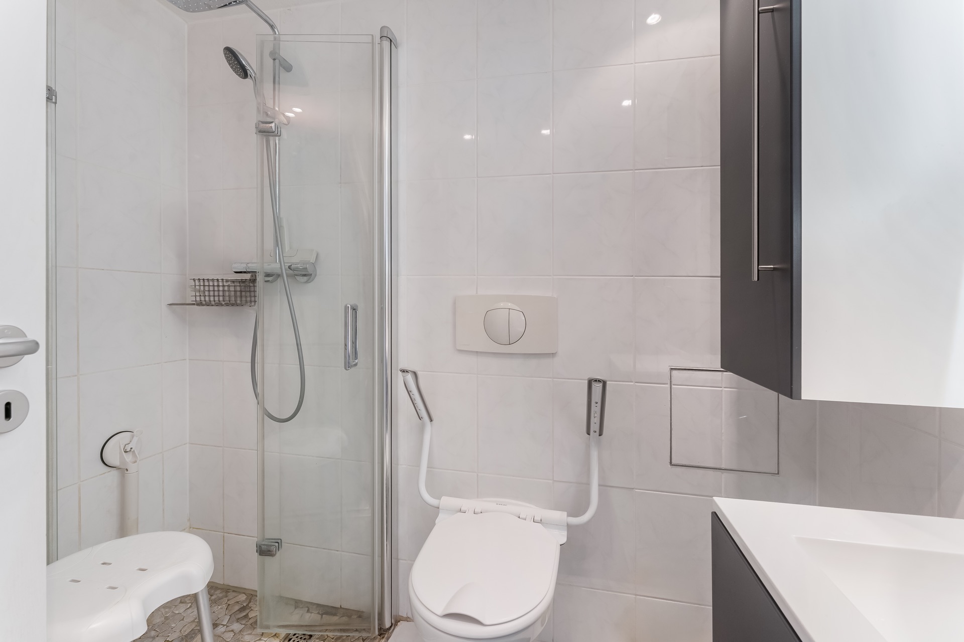 Barrierefreies Badezimmer mit Dusche, Duschhocker, Haltegriffen und Toilette.