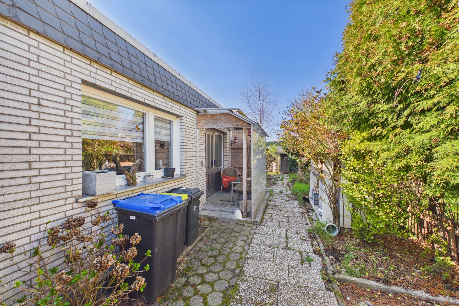 Gepflasterter Weg neben einem hellen Wohnhaus mit Mülltonnen und Gartenpflanzen.