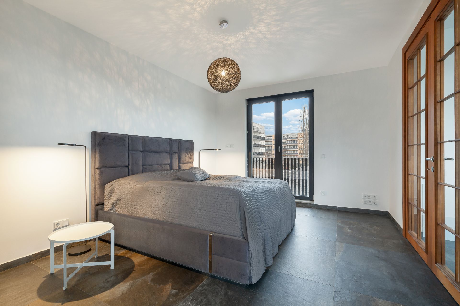 Modernes Schlafzimmer mit grauem Bett, Stehlampen, Fenstertüren und großem Fenster.
