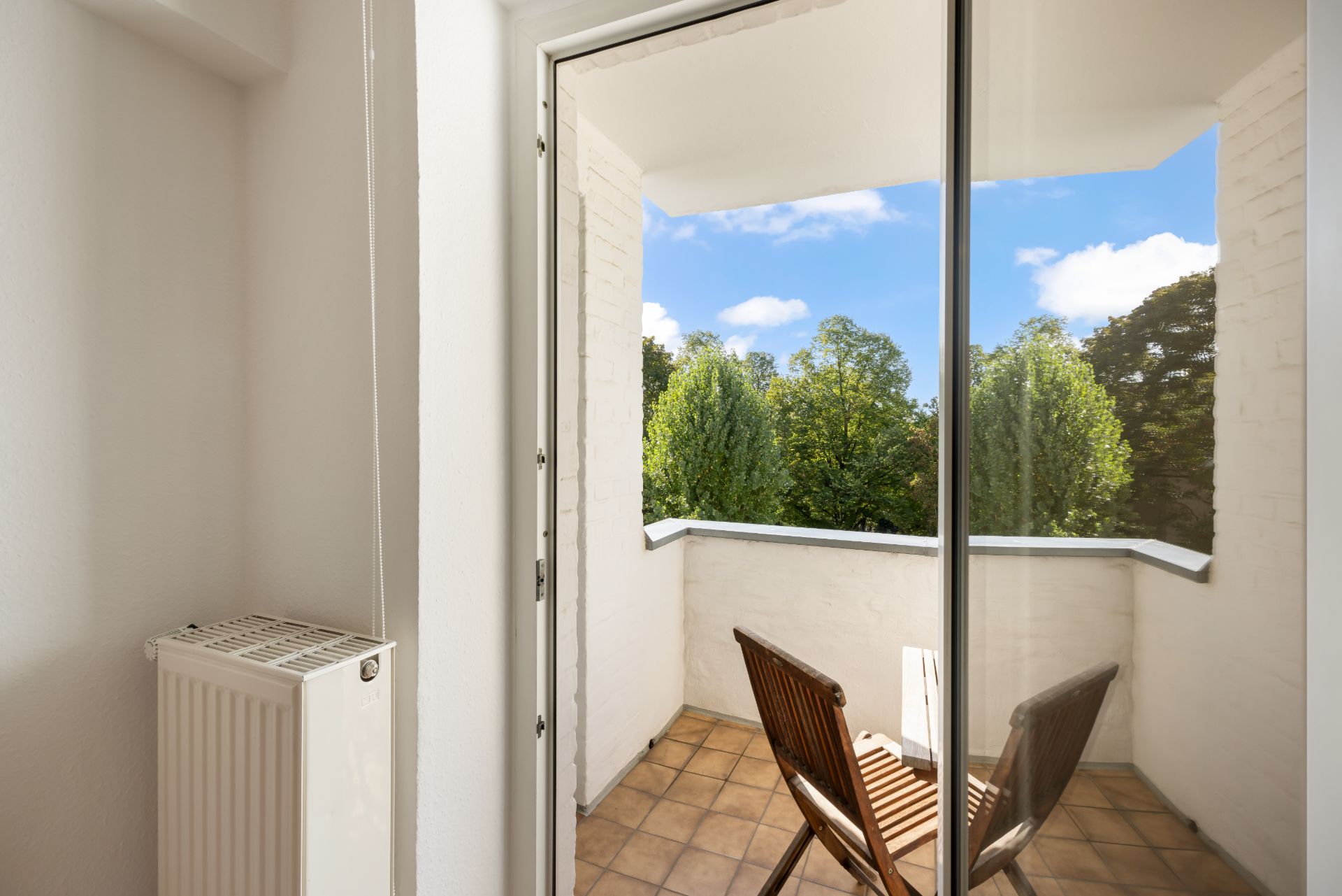 Balkon mit zwei Holzstühlen und Tisch, Blick auf grüne Bäume und blauen Himmel.