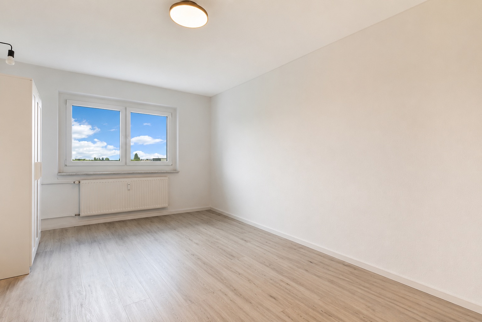 Helles, leeres Zimmer mit zwei Fenstern, Heizkörper und hellem Holzfußboden.