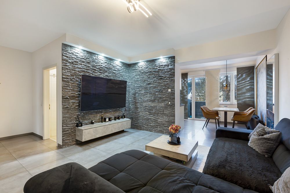 Modernes Wohnzimmer mit dunklem Sofa, TV an Steinwand und Esstisch mit Stühlen im Hintergrund.