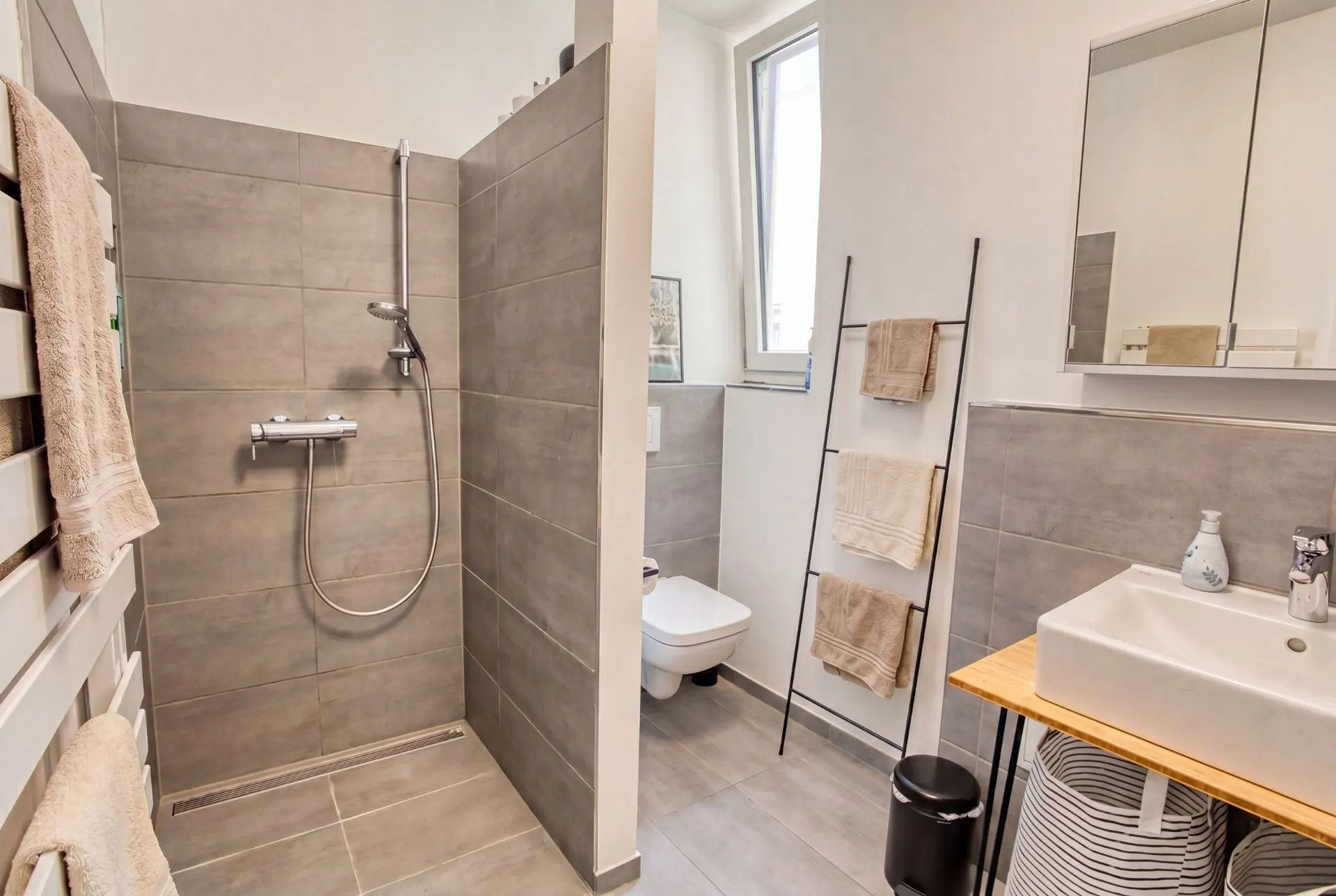 Modernes Badezimmer mit bodengleicher Dusche, WC, Waschbecken, Handtuchleiter und Fenster.