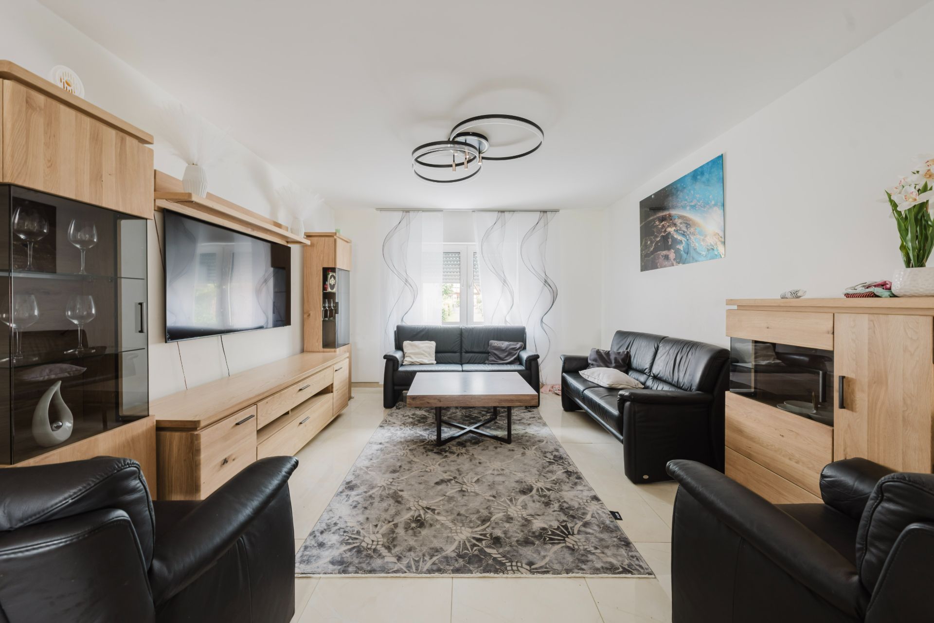 Helles Wohnzimmer mit schwarzen Ledersofas, Holzmöbeln, Teppich und großem Fenster.