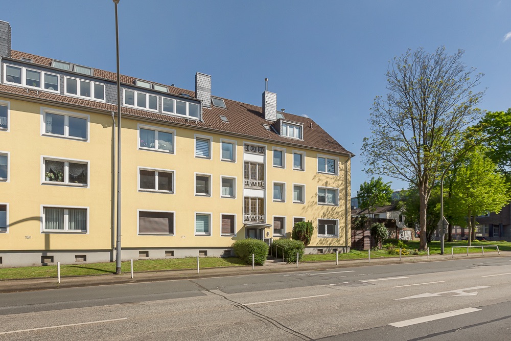 Mehrfamilienhaus mit gelber Fassade, mehreren Fenstern und Vorgarten an einer Straße.