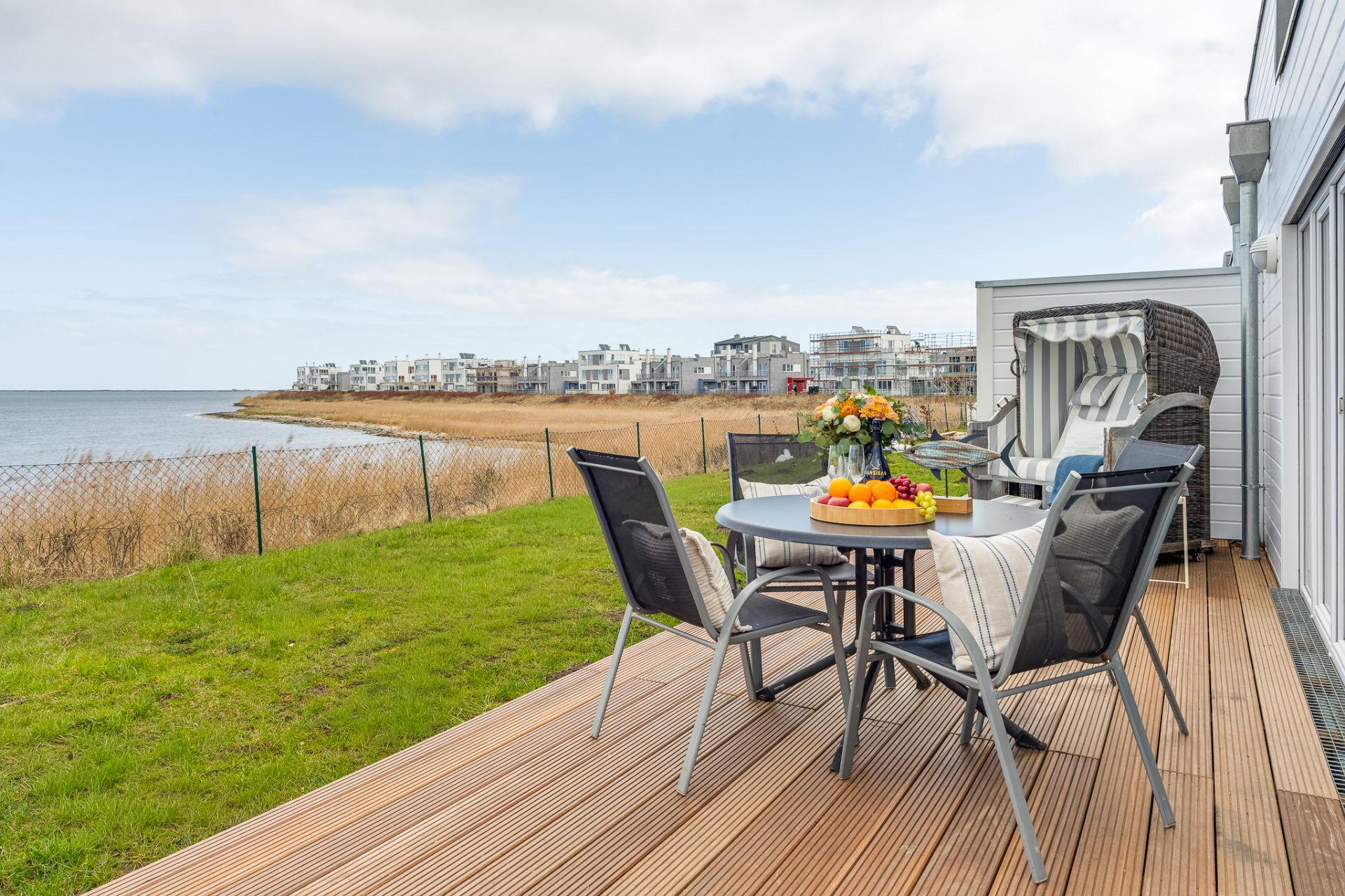 Holzterrasse mit Gartentisch, Stühlen und Strandkorb, Blick auf Wasser und Wohnhäuser.