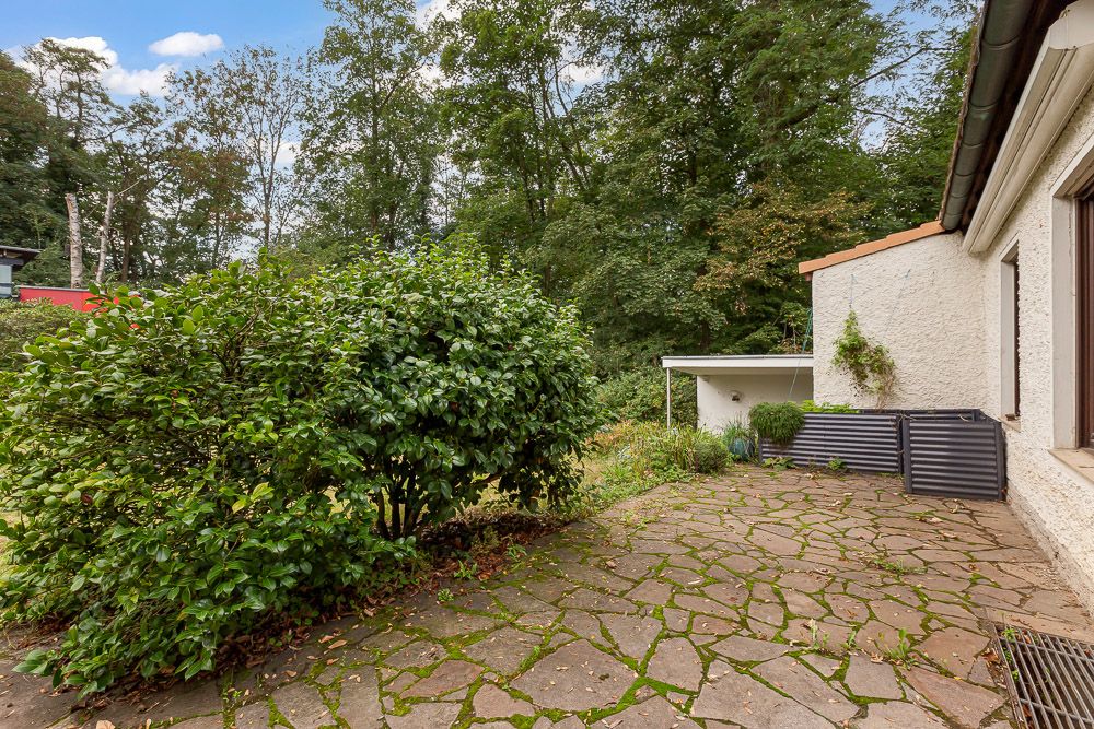 Steinplatte Terrasse mit Büschen und Blick auf einen bewaldeten Garten hinter einem Wohnhaus.