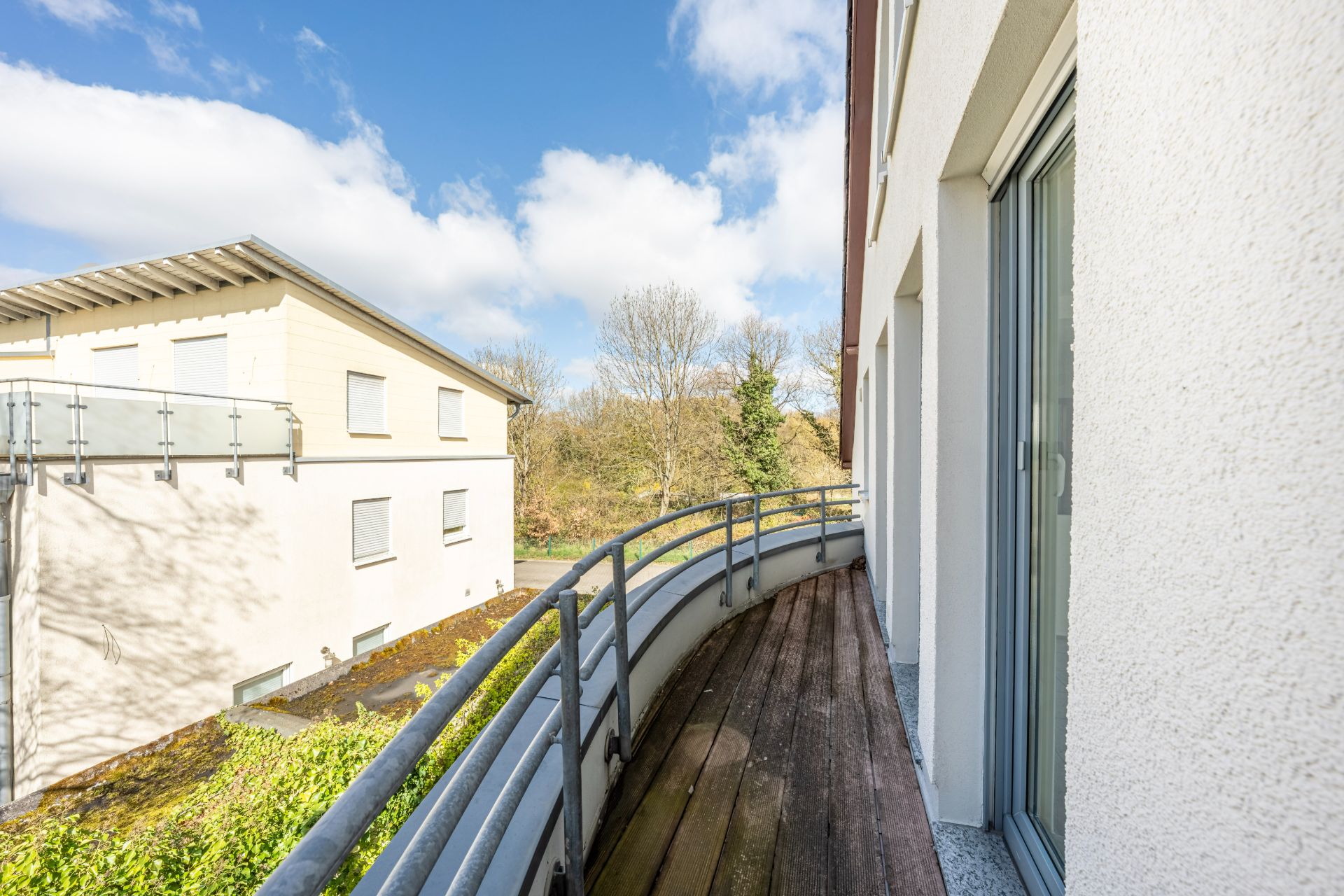 Balkon mit Holzboden und Metallgeländer, Blick auf Nachbarhaus und Bäume im Hintergrund.