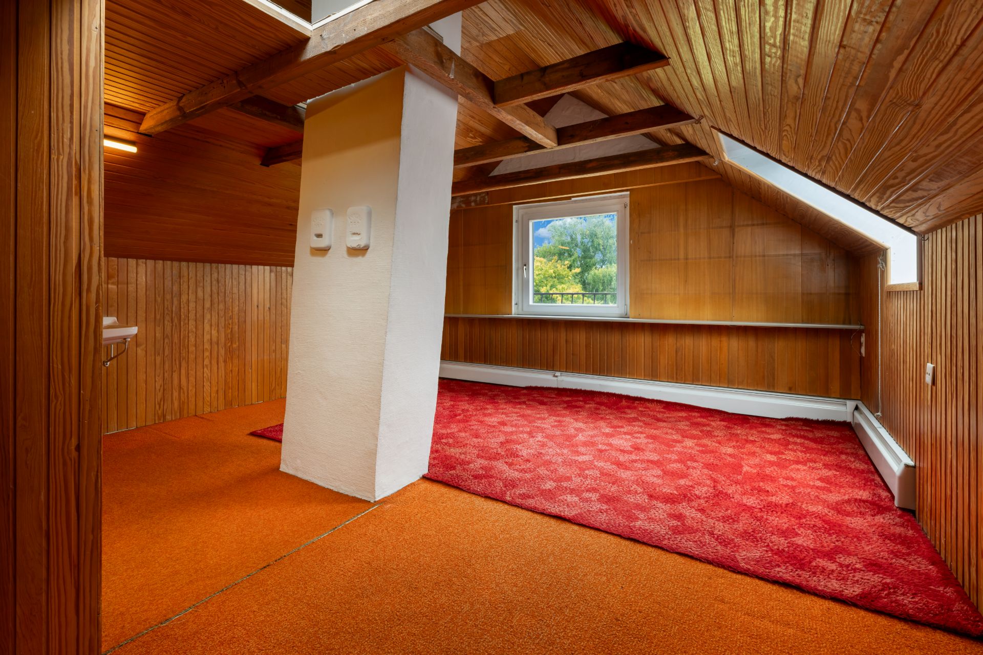 Dachgeschosszimmer mit Holzverkleidung, rotem Teppich und Fenster mit Blick nach draußen.