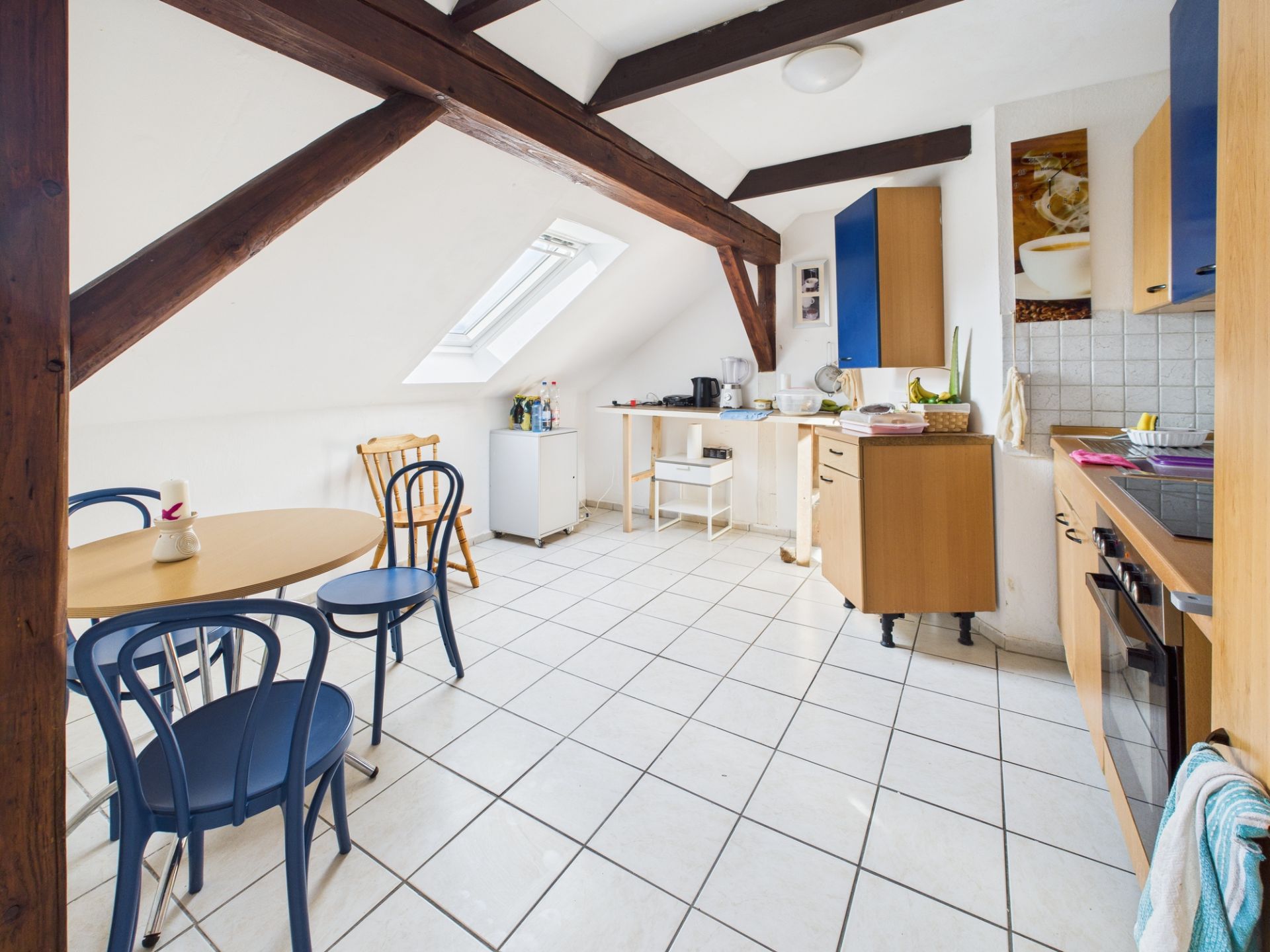Helle Küche mit Esstisch, blauen Stühlen und Dachfenster, sichtbare Holzbalken an der Decke.