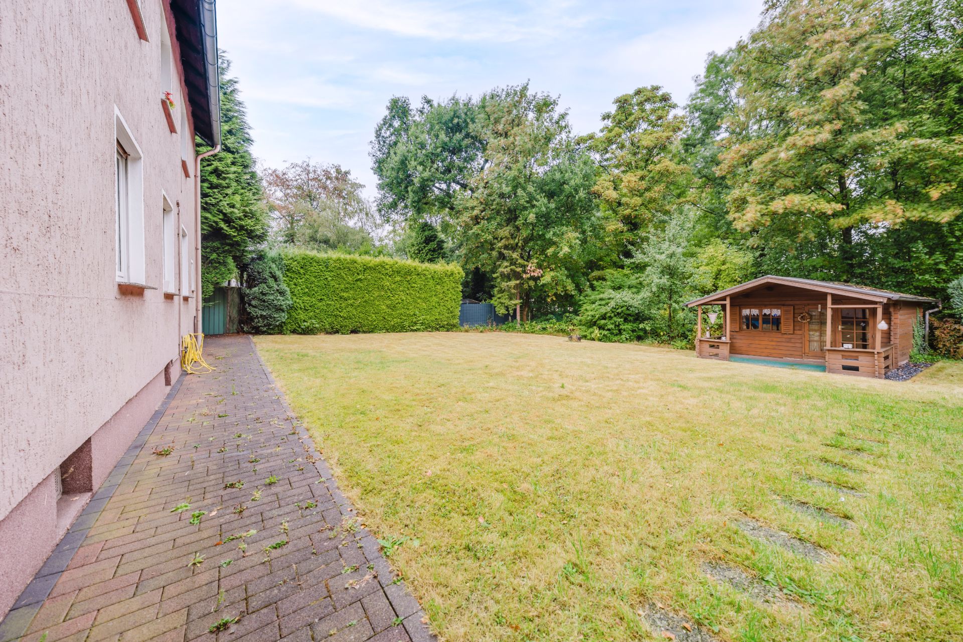 Rasenfläche mit Gartenhaus, rechts eine Hecke und viele Bäume, links Hauswand mit Fenster und Weg.