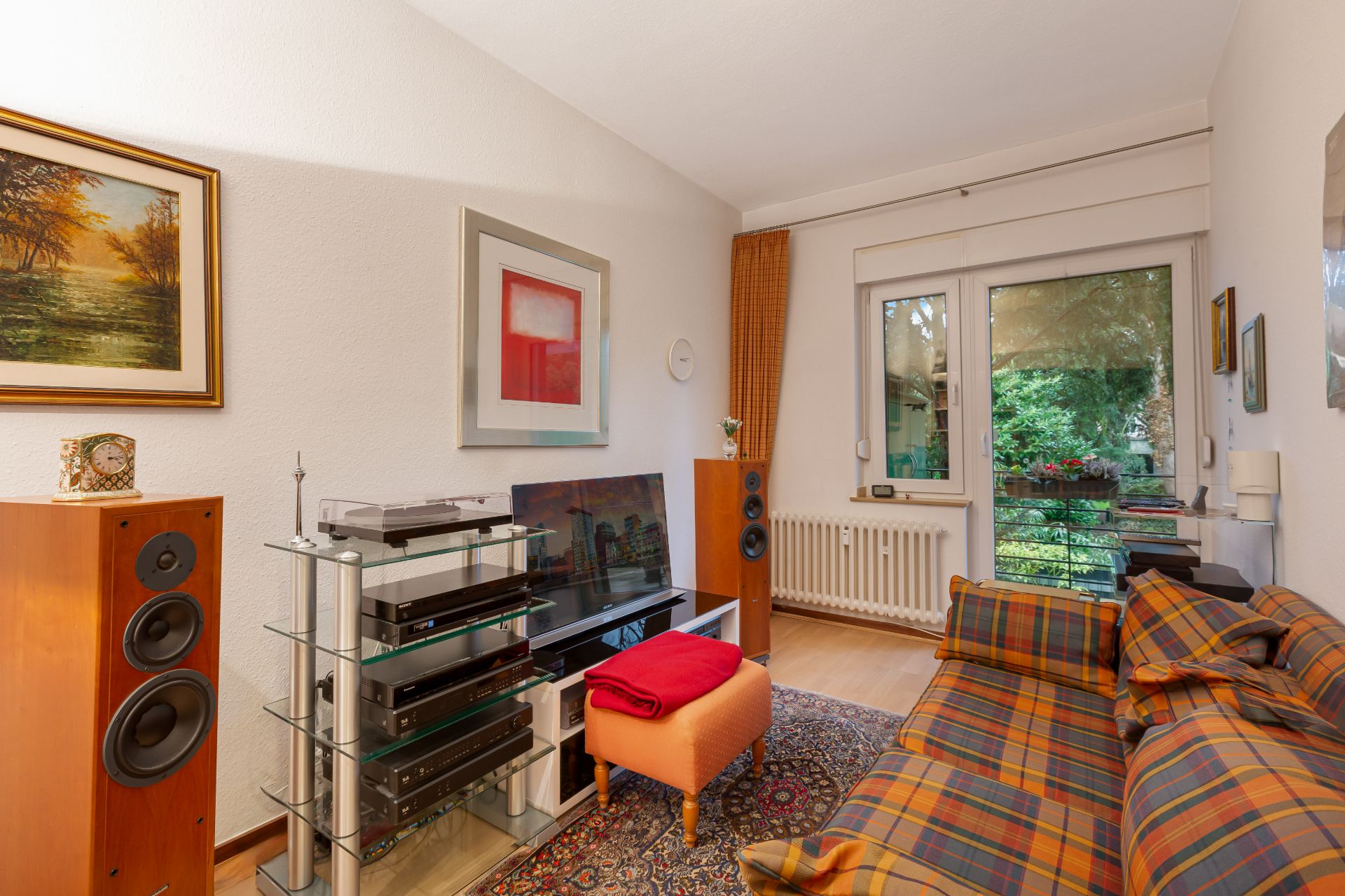 Wohnzimmer mit kariertem Sofa, Fernseher, Lautsprechern und Zugang zum Garten durch eine Glastür.