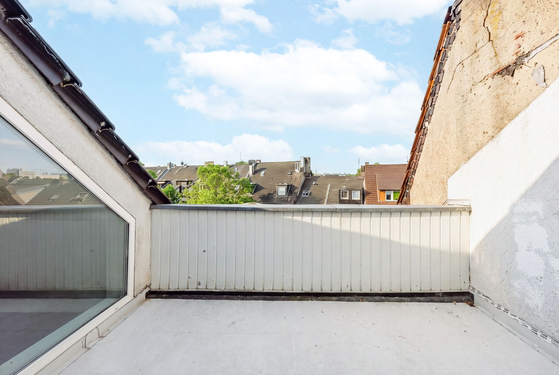 Kleine Dachterrasse mit weißer Umrandung und Blick auf Nachbarhäuser bei blauem Himmel.