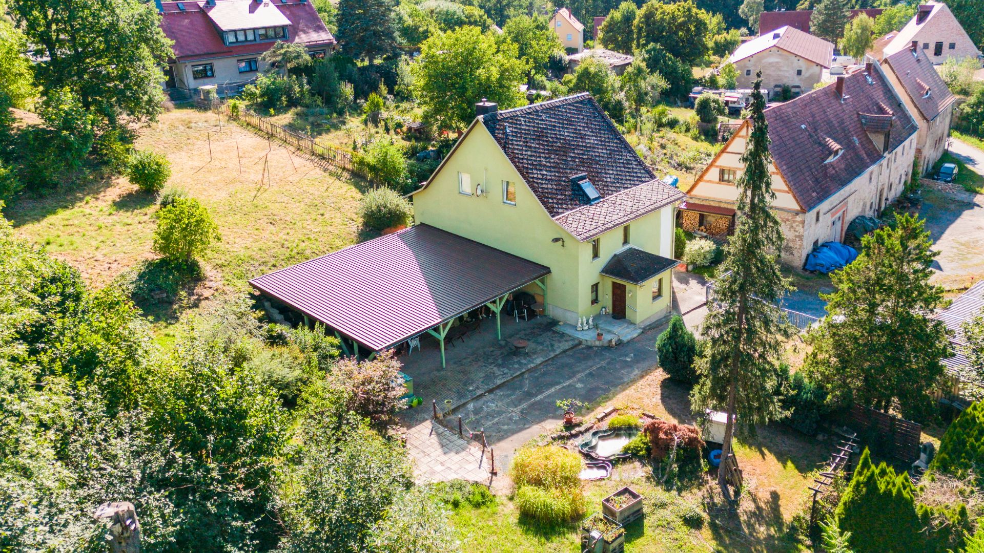 Luftaufnahme eines gelben Hauses mit Carport, Garten und umliegenden Bäumen.