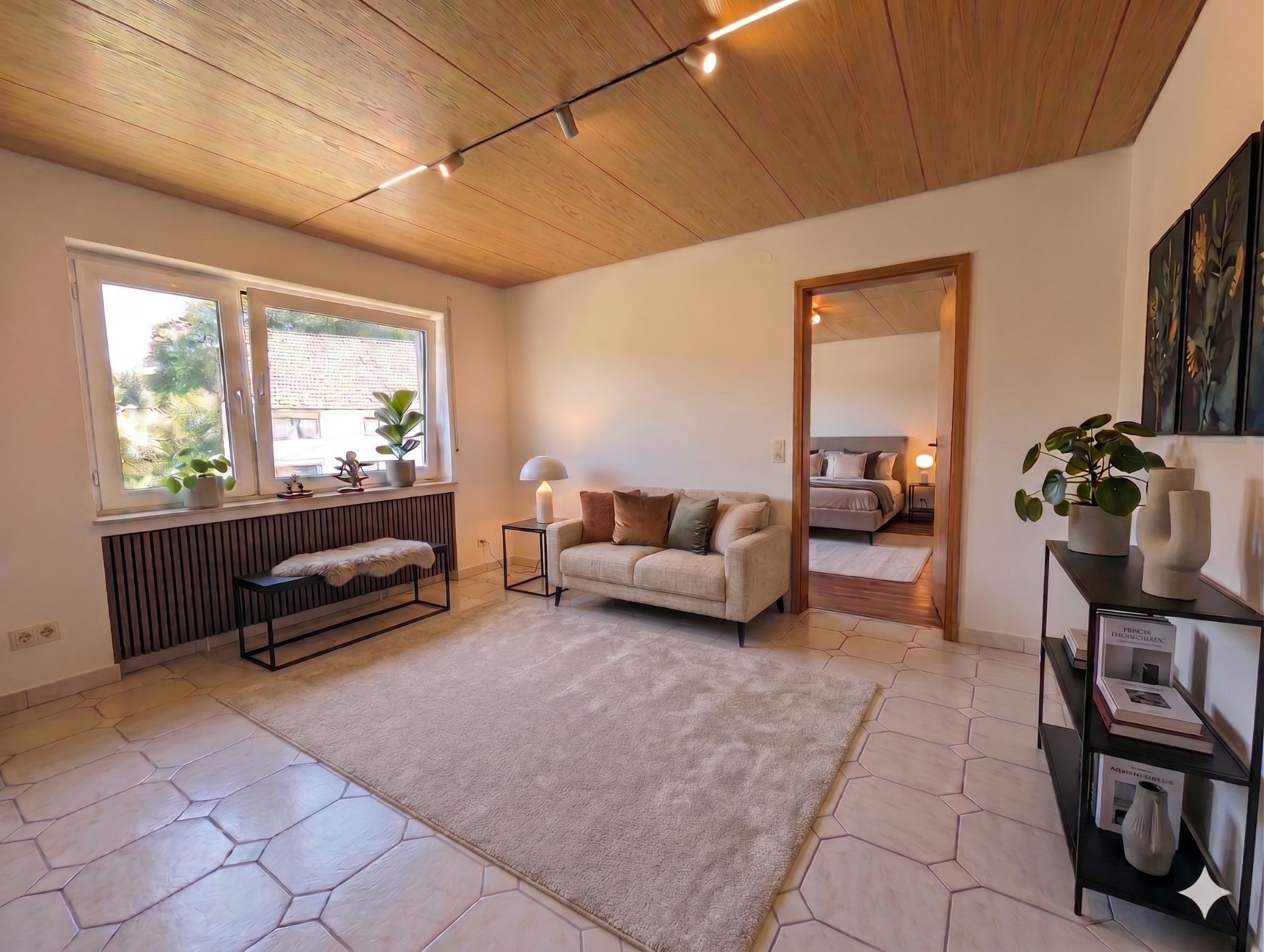 Helles Wohnzimmer mit Sofa, Teppich, Pflanzen, Regal und Zugang zu einem Schlafzimmer.