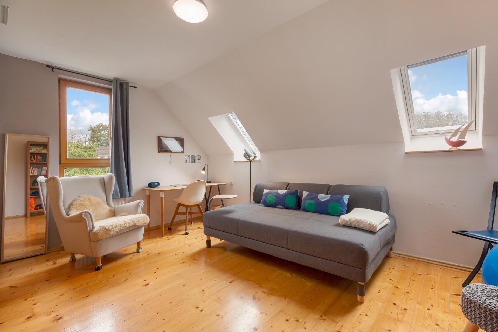 Helles Zimmer mit Sessel, Schreibtisch und grauem Sofa unter Dachschrägen.