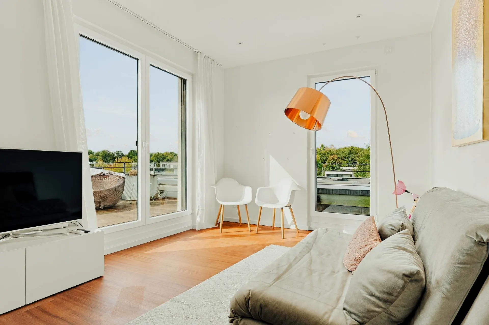 Helles Wohnzimmer mit Sofa, TV, Stehlampe und Zugang zum Balkon.