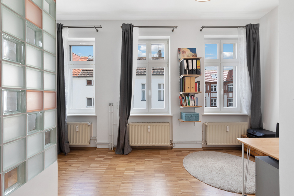 Helles Zimmer mit drei Fenstern, Vorhängen, Regal, Tisch und Parkettboden.
