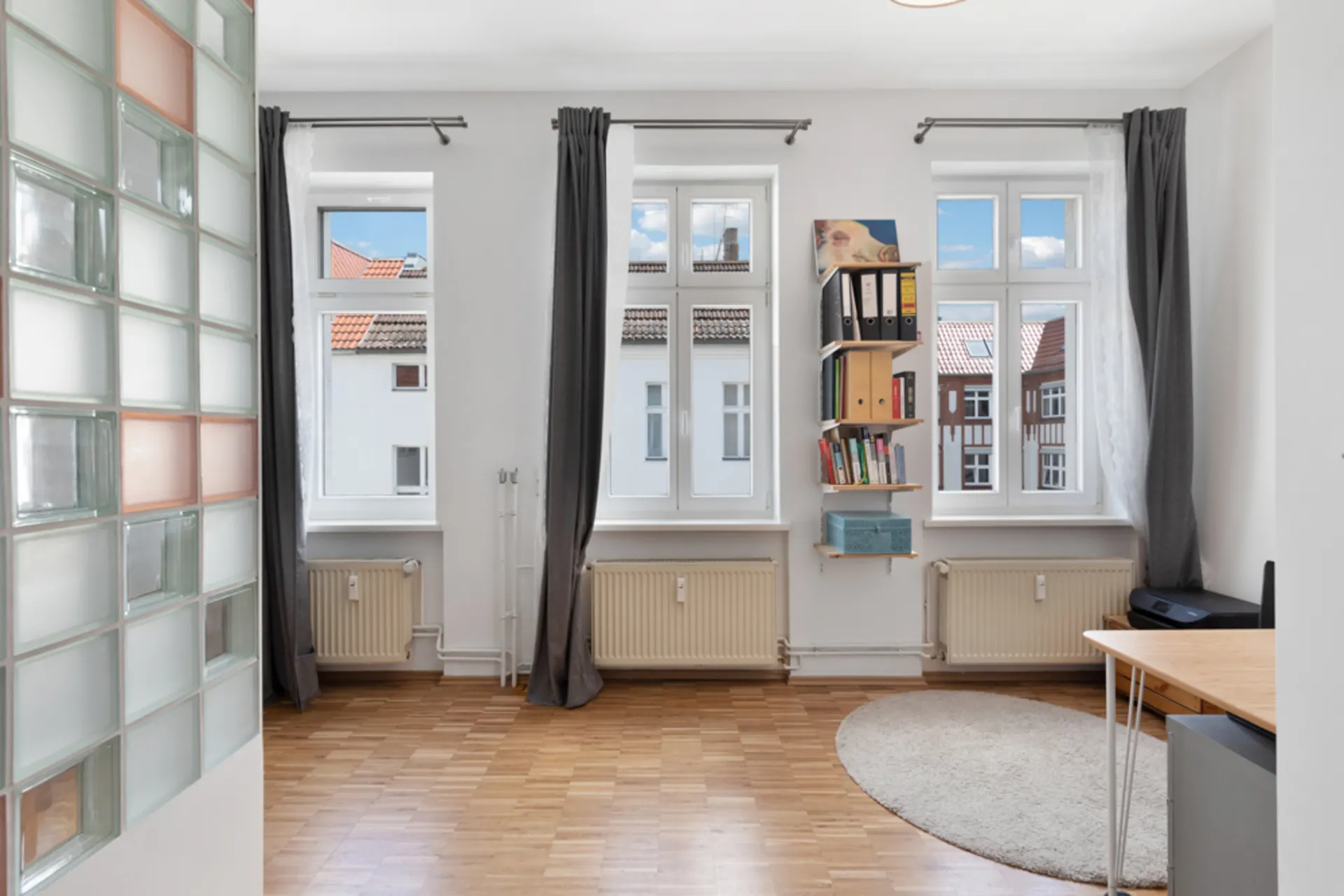Helles Zimmer mit drei Fenstern, Vorhängen, Regal, Tisch und Parkettboden.