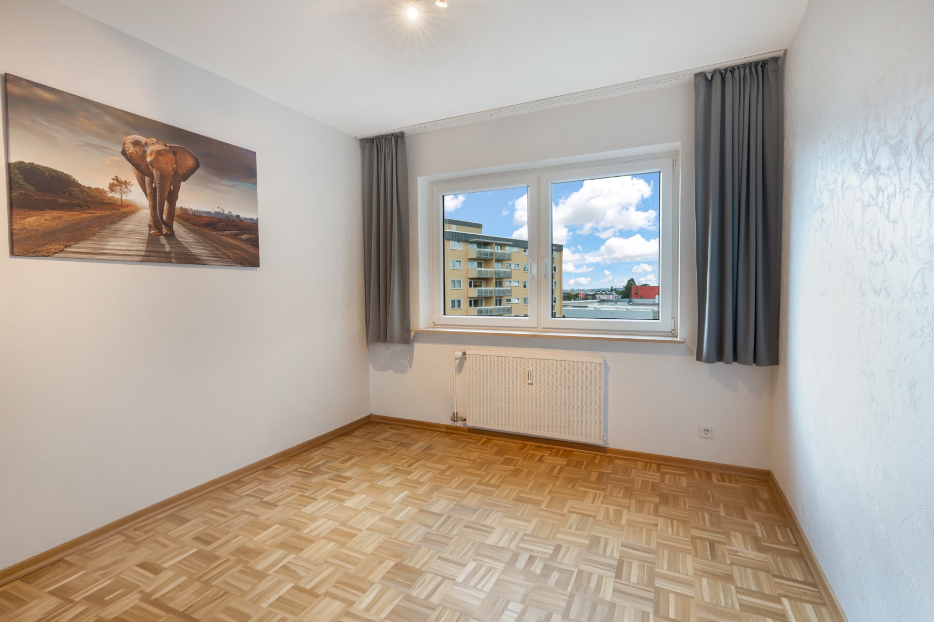 Helles, leeres Zimmer mit Parkettboden, Fenster, grauen Vorhängen und Bild von einem Elefanten an der Wand.