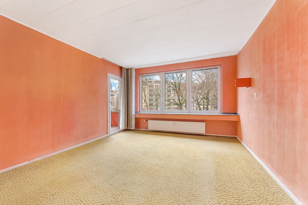 Leeres Zimmer mit orangefarbenen Wänden, großem Fenster und gelbem Teppichboden.