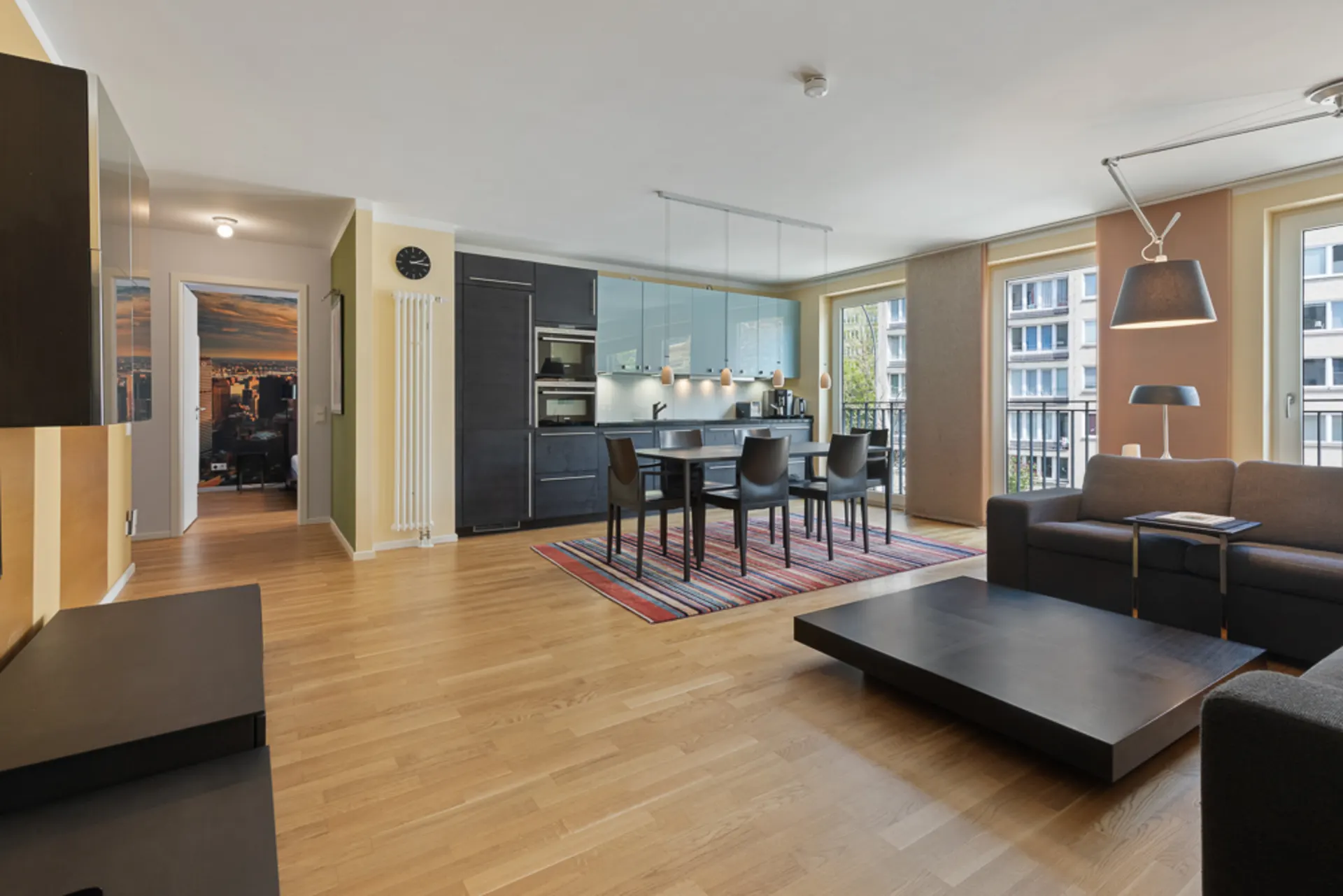 Modernes Wohnzimmer mit offener Küche, Esstisch, Sofa und großen Fenstern.