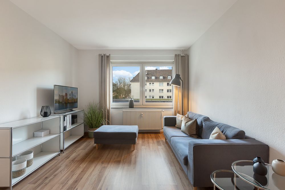 Helles Wohnzimmer mit grauem Sofa, Hocker, Regal, Fernseher und großem Fenster.