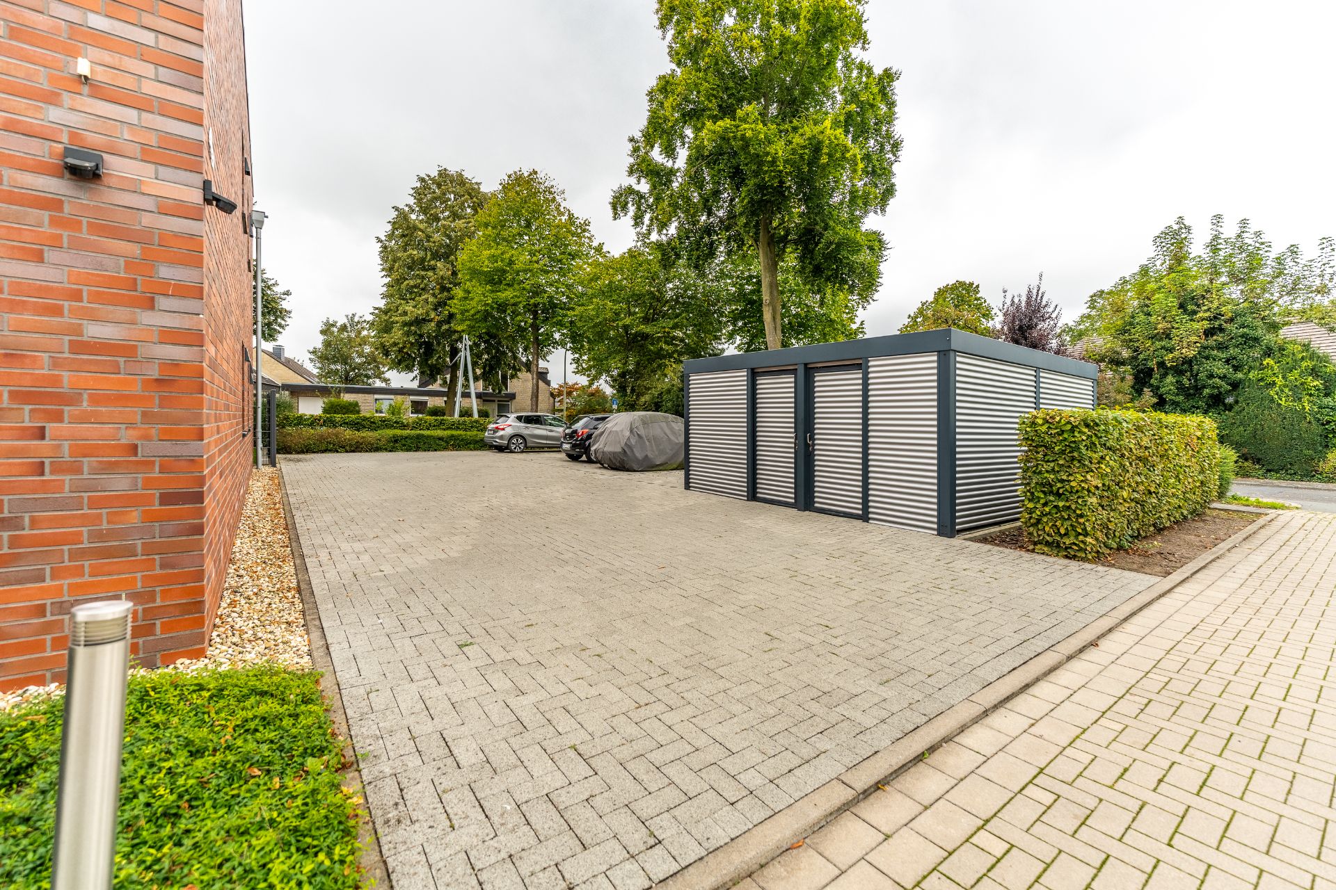 Gepflasterter Parkplatz mit moderner Metallhütte und mehreren geparkten Autos neben Bäumen.