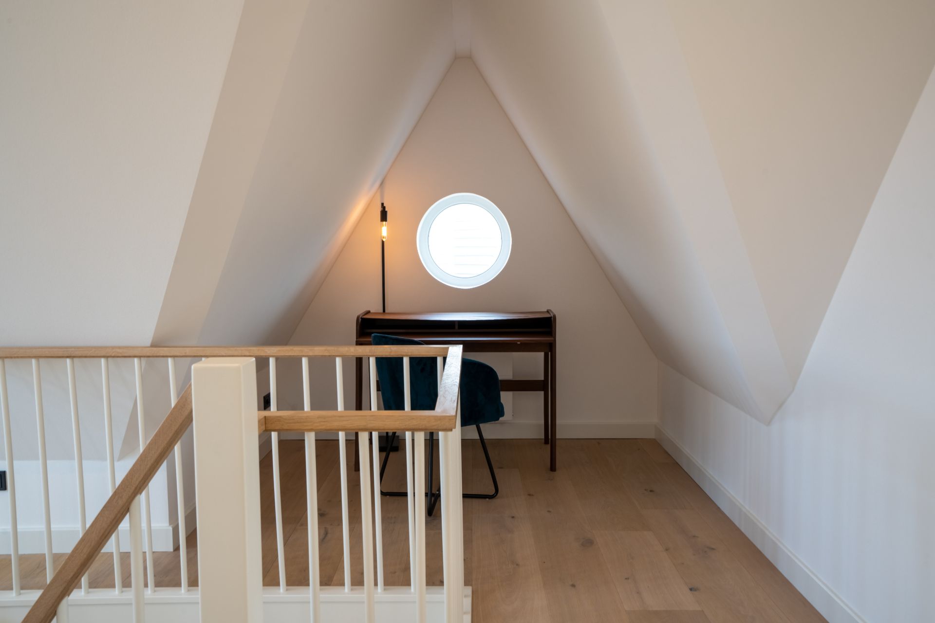 Kleiner Raum mit Schreibtisch, Stuhl, Stehlampe und rundem Fenster, sichtbar von einer Treppe.