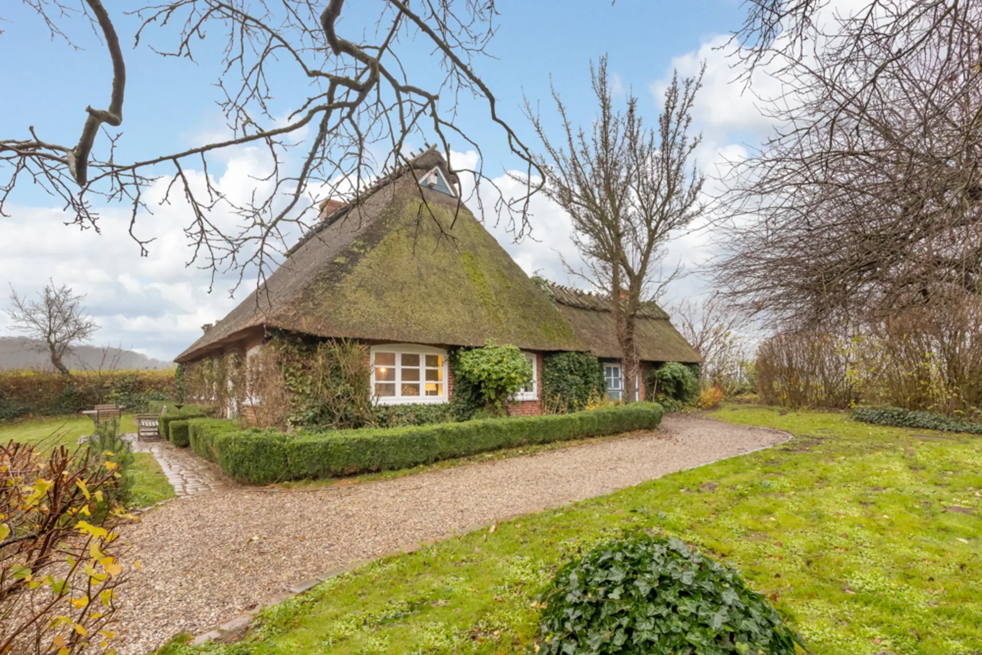Reetgedecktes Haus mit Ziegelwänden, Garten, Kiesweg und Bäumen im Vordergrund.