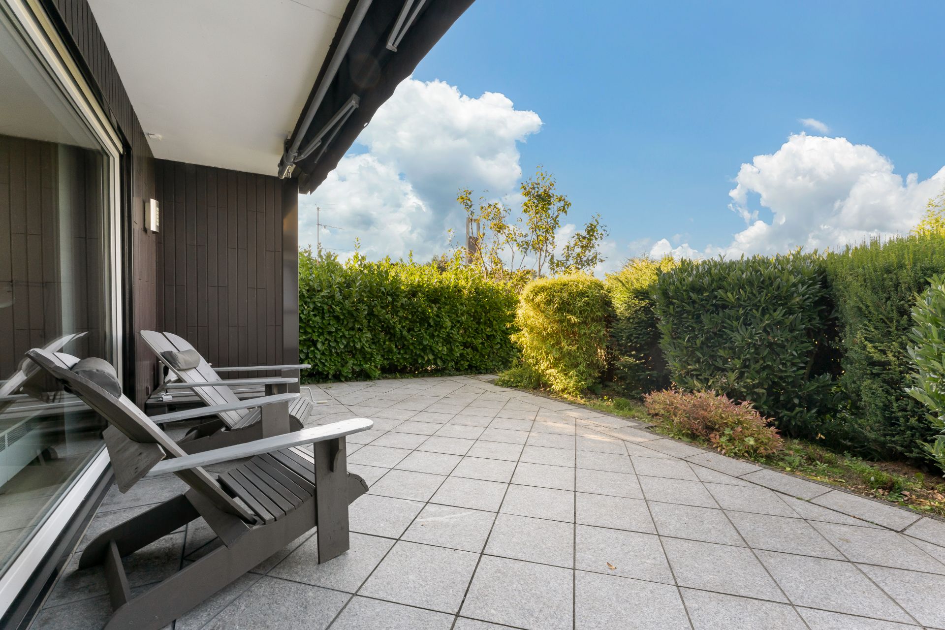 Große Terrasse mit zwei Holzstühlen und Blick auf Gartenhecken bei Sonnenschein.