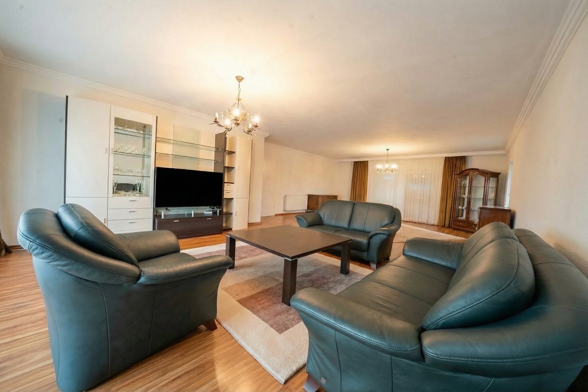 Wohnzimmer mit drei Ledersofas, Holztisch, Teppich, Schrankwand und Fernseher auf Holzfußboden.