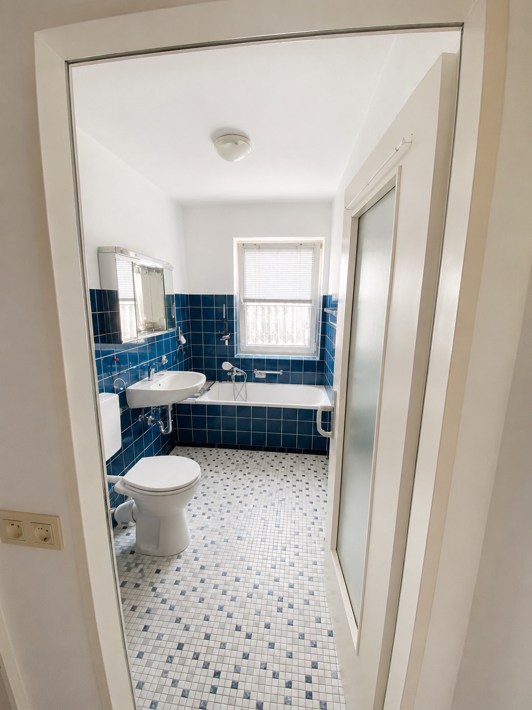 Badezimmer mit blauen Fliesen, Toilette, Waschbecken, Badewanne und Fenster.