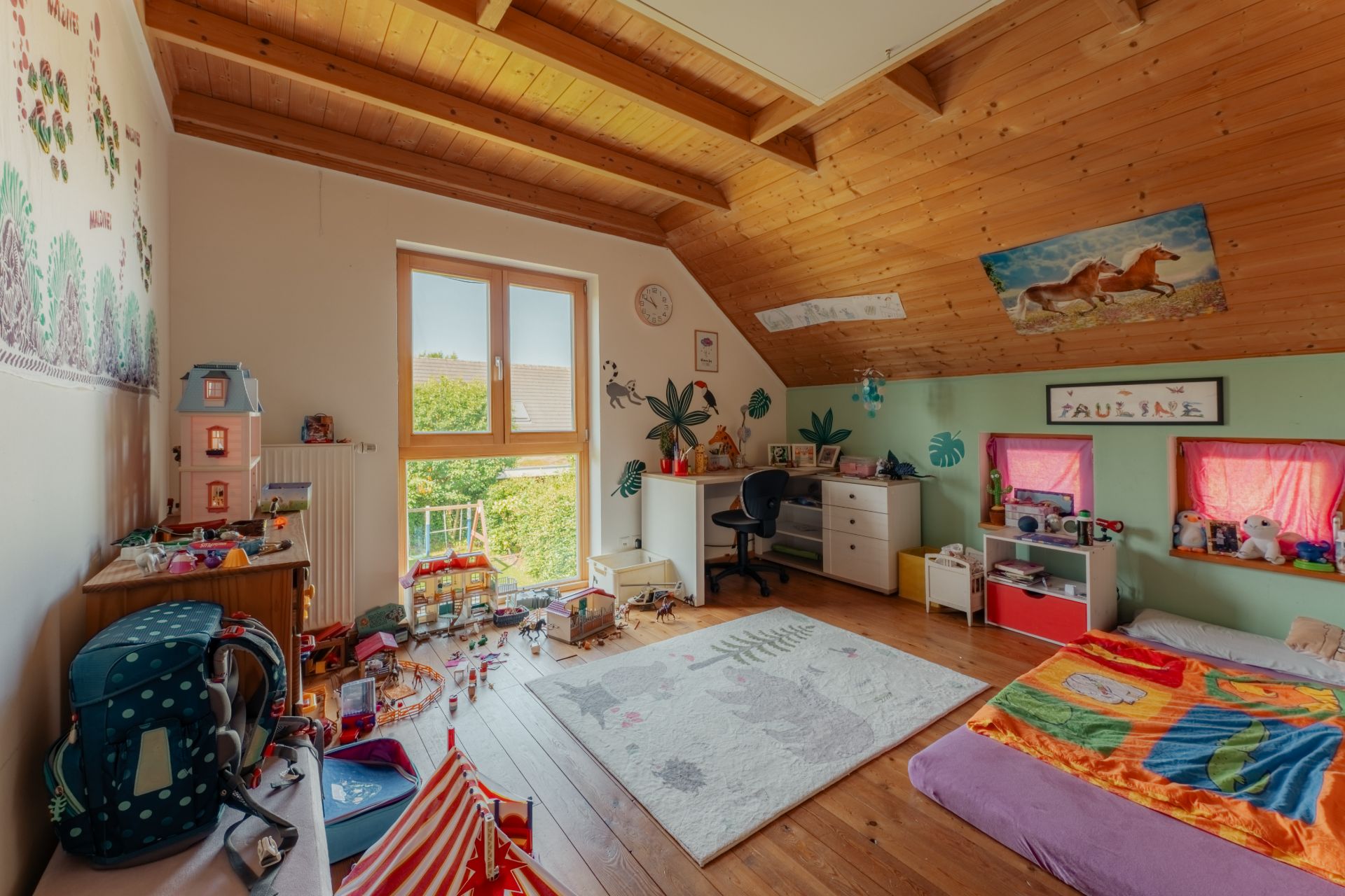 Kinderzimmer mit Holzdecke, Spielzeug, Schreibtisch, Teppich und großem Fenster mit Blick ins Grüne.