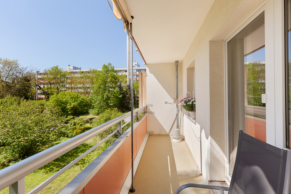 Langer Balkon mit Geländer, Blick ins Grüne und auf Wohngebäude, Sonnenmarkise oben.