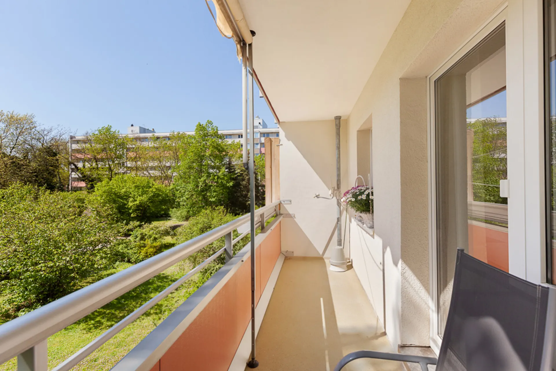 Langer Balkon mit Geländer, Blick ins Grüne und auf Wohngebäude, Sonnenmarkise oben.