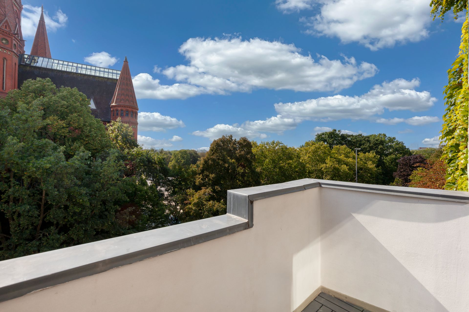 Kleine Terrasse mit Blick auf Bäume und Gebäudeturm, blauer Himmel mit Wolken.